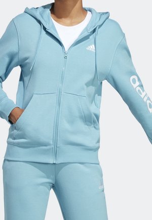 Personne portant un sweat à capuche zippé Adidas bleu clair avec poches avant et pantalon de survêtement assorti, t-shirt blanc en dessous, main dans la poche.