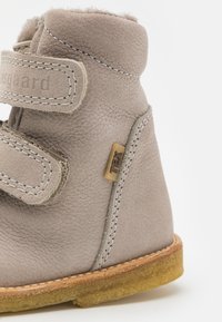 Bottine en cuir gris clair avec deux bandes Velcro, surface texturée et semelle en caoutchouc beige. Présente une petite étiquette de marque sur le côté.