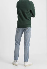 Sudadera verde, jeans azules y zapatillas blancas. La sudadera tiene un dobladillo y puños acanalados, mientras que los jeans tienen un corte holgado.