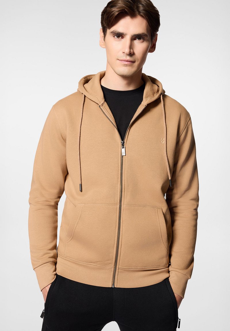 Beżowy hoodie na zamek, wykonany z miękkiej, teksturowanej tkaniny, z przednią kieszenią, regulowanym kapturem na sznurki i czarnymi akcentami na zamku.