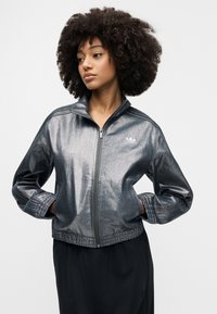 Metalna srebrna jakna na patentni zatvarač s visokim ovratnikom, rebrastim manžetama i elastičnim rubom, s Adidas logom na lijevoj strani prsa.