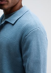 Himmelblaues, geripptes Strick-Polohemd mit klassischem Kragen und texturiertem Stoff. Das Bild konzentriert sich auf die Schulter- und ÄrmelDETAILS.