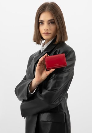 Vrouw met bruin haar en een zwarte leren jas die een rood leren portemonnee vasthoudt tegen een witte achtergrond.