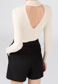 Cremfarbener Strickpullover mit tiefem V-Ausschnitt auf der Rückseite, mit enganliegenden langen Ärmeln. Kombiniert mit schwarzen Hochbundshorts mit seitlichen Taschen.