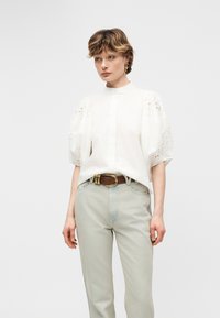 Γυναίκα με λευκό πουκάμισο με φουσκωτά μανίκια από δαντέλα με οπές, μπεζ τζιν ψηλόμεσο, καφέ ζώνη και χρυσά σκουλαρίκια, στέκεται μπροστά σε λευκό φόντο.