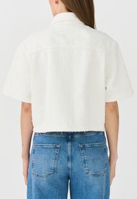 Cropped white textured jacket with short sleeves, raw hem, and collared neckline, paired with blue denim jeans featuring back pockets.