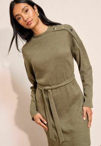 Olivza zaļa ribota adīta kleita ar augstu kaklu, garām piedurknēm un jostu viduklī. Iezīmēta ar pogām uz pleca un mīkstu tekstūru.