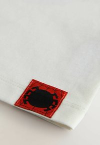 Witte textiel met een rood stuk stof met een spinontwerp en zwarte details. Het stuk is op de zoom van het shirt gestikt.