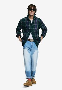 kariertes Flanellhemd in Dunkelgrün mit lila Akzenten, kombiniert mit hellblauen Jeanshosen mit Patchwork. Braune Wildlederschuhe runden den Look ab.