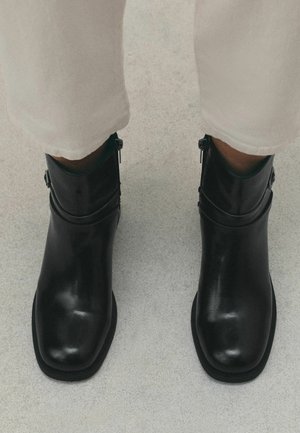 Person wearing black leather ankle boots with zippers and buckles, paired with light-colored pants on a textured floor.