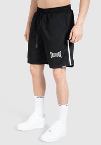 Svarta shorts med ribbad textur och vitt "SMILODOX"-logotyp. Har en dragsko i midjan, vita sidostripes och en avslappnad passform.