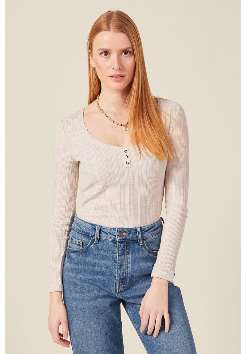 Langärmliges, beiges, geripptes Oberteil mit Rundhalsausschnitt, das vorne mit drei Knöpfen versehen ist, kombiniert mit hochgeschnittenen blauen Jeans.