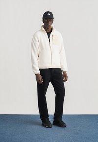 Άνδρας που φοράει μπεζ φλις μπουφάν με φερμουάρ, μαύρο παντελόνι, μαύρα αθλητικά παπούτσια και μαύρο καπέλο, στέκεται σε μπλε χαλί μπροστά σε λευκό τοίχο.