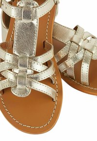 Sandales métalliques dorées avec des bretelles entrecroisées, finition texturée, semelle intérieure marron et bords cousus. Design à bout ouvert.