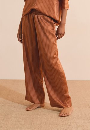 Person standing barefoot wearing loose, rust-colored striped pants and matching top against a plain beige background.