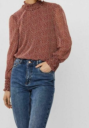 Femme portant un chemisier couleur rouille à col haut et manches longues avec des pois blancs et un jean bleu taille haute, main dans la poche, sur fond uni.