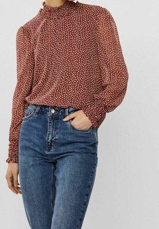 Femme portant un chemisier couleur rouille à col haut et manches longues avec des pois blancs et un jean bleu taille haute, main dans la poche, sur fond uni.