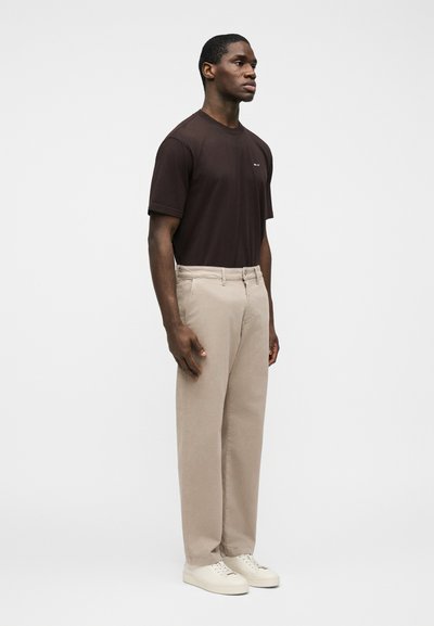 Homme debout portant un t-shirt à manches courtes marron foncé, un pantalon beige et des baskets blanches, sur un fond clair uni.