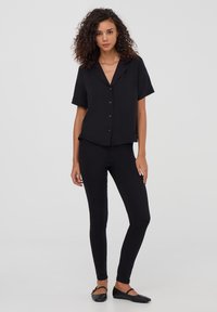 Camicia nera a maniche corte con bottoni abbinata a leggings neri aderenti e scarpe nere senza tacco, con un colletto semplice e una texture liscia.