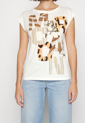 Kobieta w białym bezrękawnym topie z dużym, mieszanym zwierzęcym nadrukiem i paskowanymi literami, w połączeniu z jasnoniebieskimi dżinsami.