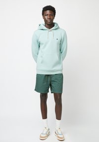 Felpa leggera verde menta con cappuccio e tasca a marsupio, abbinata a pantaloni corti verde scuro. Calze bianche e scarpe beige completano il look.