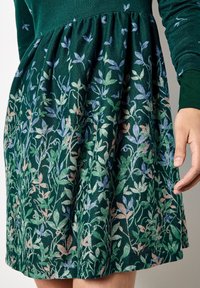 Abito verde con un tessuto testurizzato caratterizzato da un motivo floreale in colori pastello, gonna plissettata e maniche lunghe. Mano visibile all'orlo.