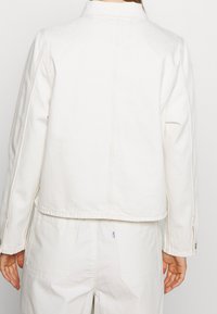 Vit denimjacka med en kortad passform, synliga sömmar och krage med spets. Har långa ärmar och knappade manschettdetaljer.