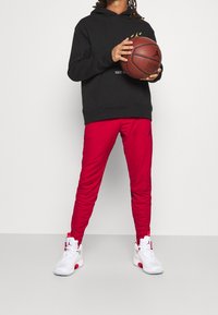 Sudadera negra con acentos amarillos, pantalones deportivos rojos y zapatillas blancas con detalles rojos, sosteniendo un balón de baloncesto en un entorno neutral.