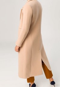 Une personne portant un long manteau en laine beige avec une fente au dos, un pantalon court brun et des chaussures noires à enfiler, sur un fond clair uni.