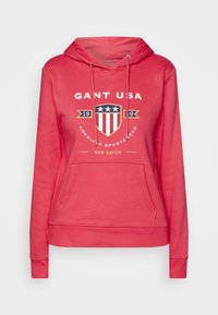 Sweat à capuche rouge avec une poche avant, capuche à cordon de serrage, et graphique imprimé représentant un bouclier avec des étoiles et des rayures. Confectionné en tissu doux.