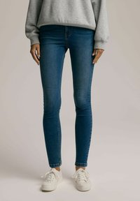 Chude jeansy w ciemnoniebieskim denimu, z wysokim stanem, dopasowanym krojem i lekkim rozciąganiem. W zestawie z białymi sneakersami.