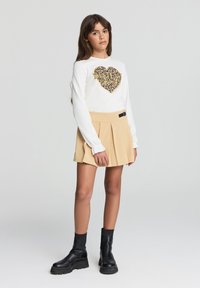 Biała koszula z długim rękawem z sercem w złotym i leopardzim wzorze, zestawiona z beżową spódnicą plisowaną i czarnymi botkami za kostkę.