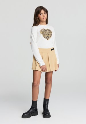 Camicia bianca a maniche lunghe con un design a cuore in oro e stampa leopardata, abbinata a una gonna plissè beige e stivaletti neri alla caviglia.