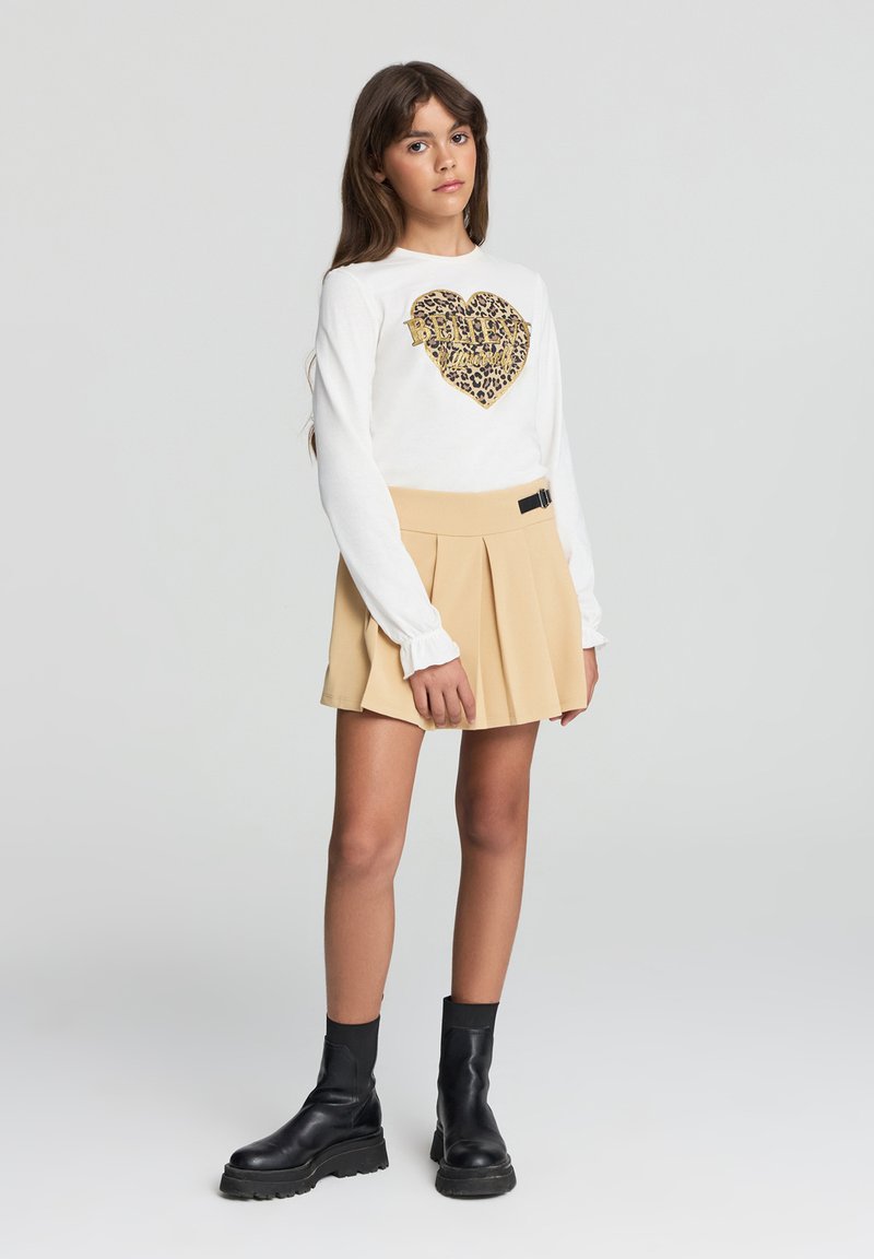 Biała koszula z długim rękawem z sercem w złotym i leopardzim wzorze, zestawiona z beżową spódnicą plisowaną i czarnymi botkami za kostkę.