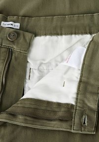 Pantaloni verde oliva con zip e chiusura a bottone, e una fodera interna a righe bianche. Etichette visibili che indicano la taglia 32/32.