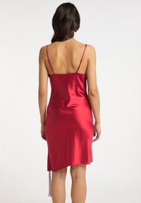 Robe nuisette en satin rouge avec des bretelles réglables, texture lisse, coupe mi-genoux et un détail de cordon sur le côté pour un ajustement personnalisable.