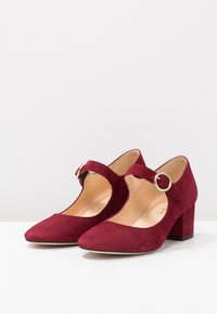 Talons en daim bordeaux avec un bout arrondi, des côtés découpés et une sangle ornée d'une boucle en métal circulaire. Talon épais pour plus de stabilité.