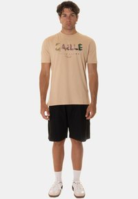 T-shirt in cotone beige con testo stampato multicolore, abbinata a pantaloni corti neri. Calzini e scarpe bianche completano l'outfit. Il modello è in piedi.