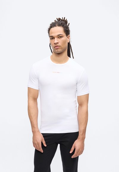 Άνδρας με ράστα, φορώντας εφαρμοστό λευκό πουκάμισο με κοντά μανίκια και μαύρο παντελόνι, σε απλό φωτεινό φόντο.