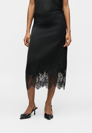 Black midi skirt with sheer lace hem detailing, worn with black pointed-toe slingback heels on a model against a plain background.