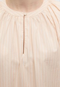 Κοντινό πλάνο κρεμ μπλούζας με λεπτές πορτοκαλί κάθετες ρίγες, στρογγυλή λαιμόκοψη και κούμπωμα με κουμπί καλυμμένο με ύφασμα στο γιακά.