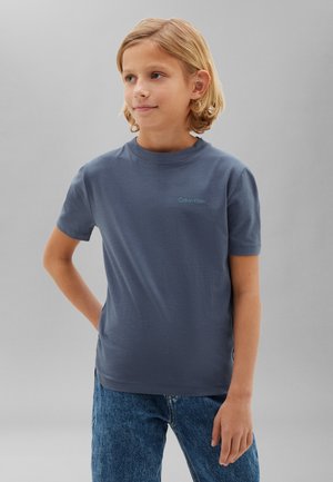 Blondes Kind mit schulterlangem Haar, das ein blaues Calvin Klein T-Shirt und blaue Jeans trägt, steht mit einer Hand hinter dem Rücken.