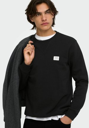 Jeune homme aux cheveux bruns mi-longs porte un sweat noir et un sous-vêtement blanc, tenant un manteau foncé sur son épaule et regardant sur le côté.