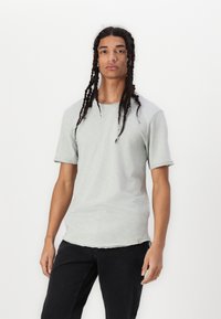 Hellgraues T-Shirt mit kurzen Ärmeln und Rundhalsausschnitt, aus weichem Stoff, mit leicht ausfransendem Saum und lässiger Passform. Schwarze Hose.