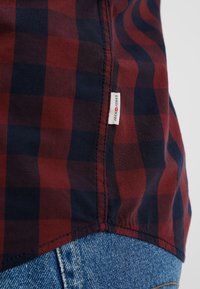 Lähedalt vaadatuna punane ja tumesinine ruuduline särk, mille hemil on "Jack & Jones" silt, siniste denim pükste kohal.