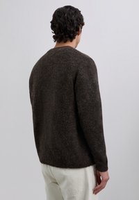 Hombre con cabello oscuro y ondulado, vestido con un suéter marrón oscuro de felpa y pantalones beige claros, de pie de espaldas contra un fondo claro y liso.