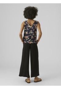 Hedvábný top bez rukávů s květinovým vzorem v námořnické modři a designem V-zad, kombinovaný s volnými černými kalhotami a béžovými sandály. Hladká struktura látky.