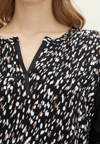 Schwarze Bluse mit V-Ausschnitt, die ein verstreutes Muster aus weißen und braunen Punkten aufweist. Leichter Stoff mit kurzen Ärmeln.