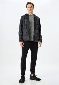 Zwarte geprinte zip-up jas over een grijs shirt, gecombineerd met zwarte slim fit broek en zwarte sportschoenen. Glad textuur, lichtgewicht materiaal.