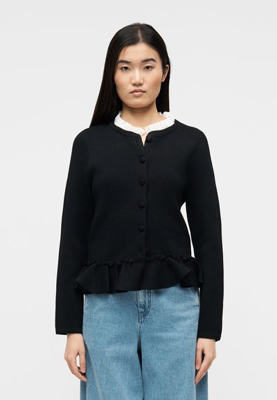 Femme portant un cardigan noir boutonné avec un ourlet à volants sur un chemisier blanc à col à volants et un jean bleu clair ample, sur un fond uni.